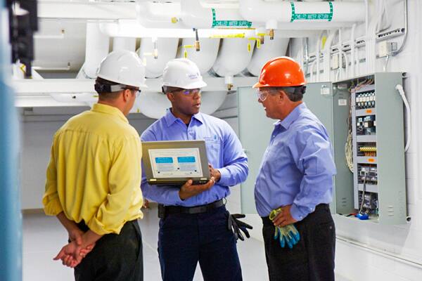 Three technicians reviewing a portable monitoring device in an industrial equipment room.